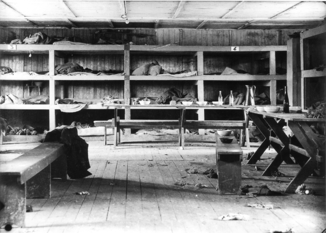 Zu sehen ist das Innere einer Häftlingsbaracke. Hinten siehjt man die vierstöckigen schlafpritschen. Im Vordergrund sind Holzbänke zu sehen. Die Baracke ist verlassen. Strohsäcke auf den Pritschen und vereinzeltes Essgeschirr zeigen von den hier untergebrachten Häftlingen.