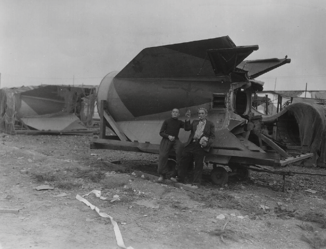 Schwarz-Weiß-Fotografie von zwei befreiten Häftlingen des KZ Mittelbau-Dora vor dem Heck einer V2-Rakete