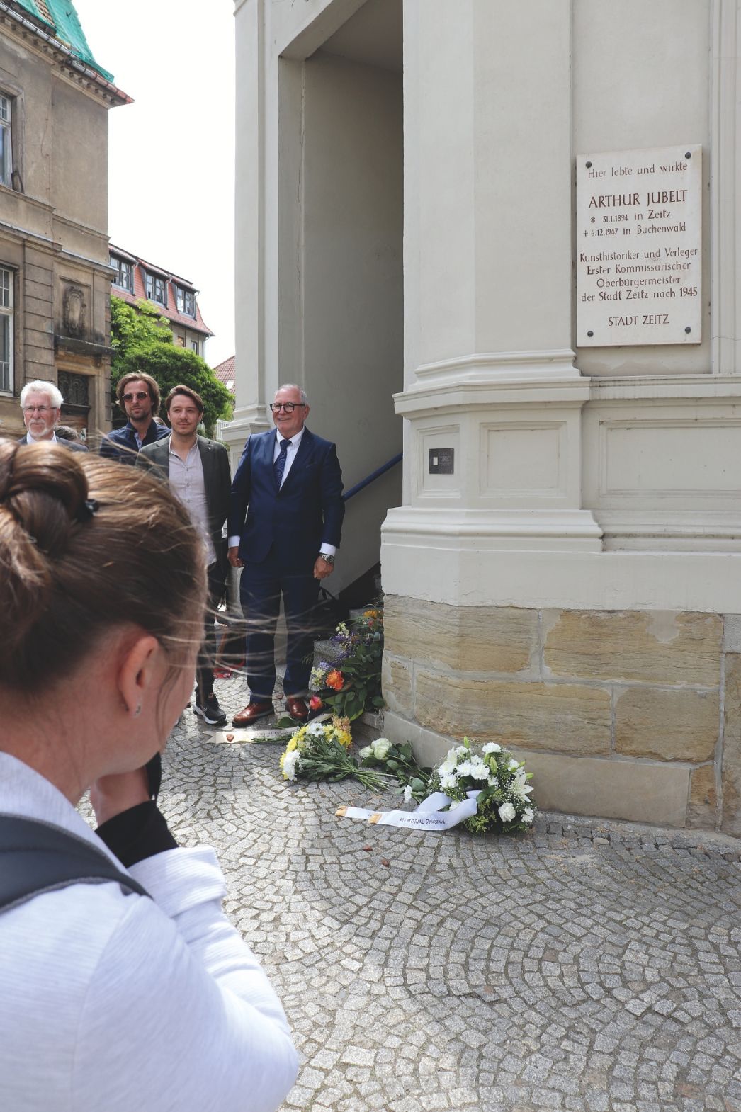 Tafel für Arthur Jubelt am ehemaligen Wohnhaus in Zeitz, davor Kränze und Besucher*innen