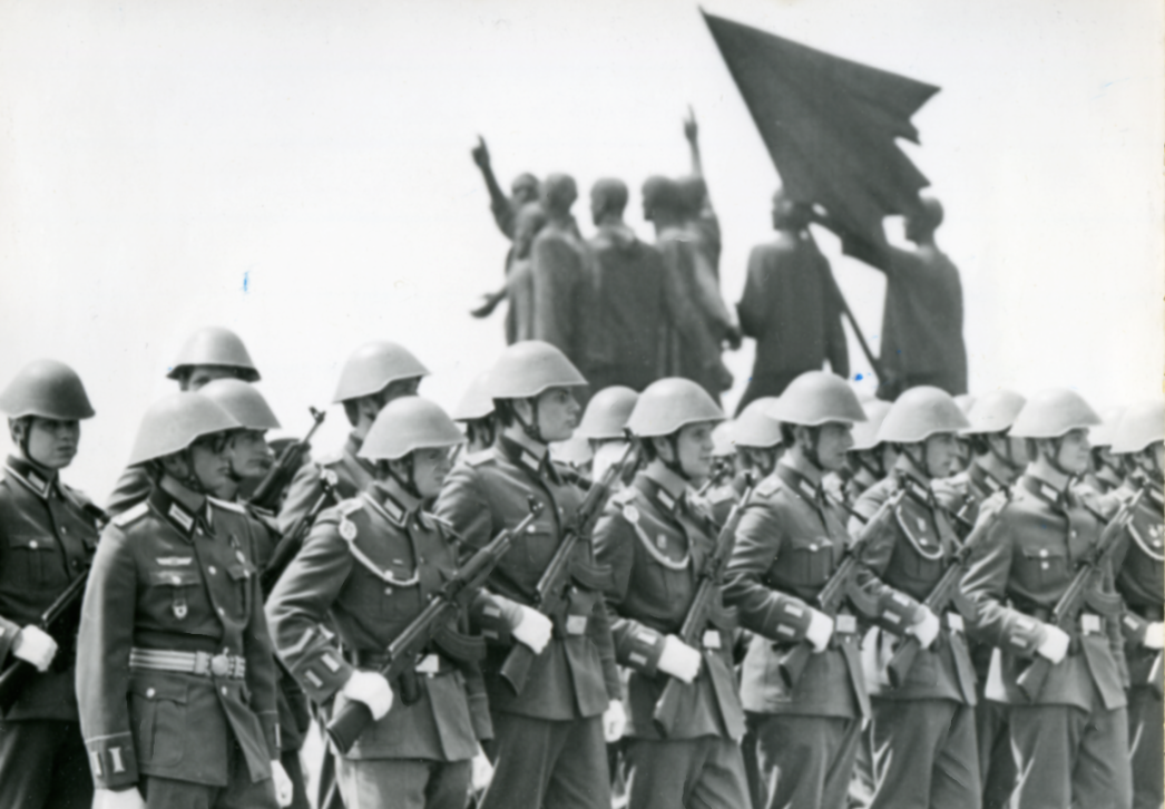 Zushen sind in Reih und Glied aufgestellte Soldaten der Nationalen Volksarmee. Im Hintergrund ist die von Fritz Cremer gestalteten Figurengruppe des Mahnmals von hinten zu sehen. Die Soldaten sind also dem Glockenturm des Mahnmals zugewandt.
