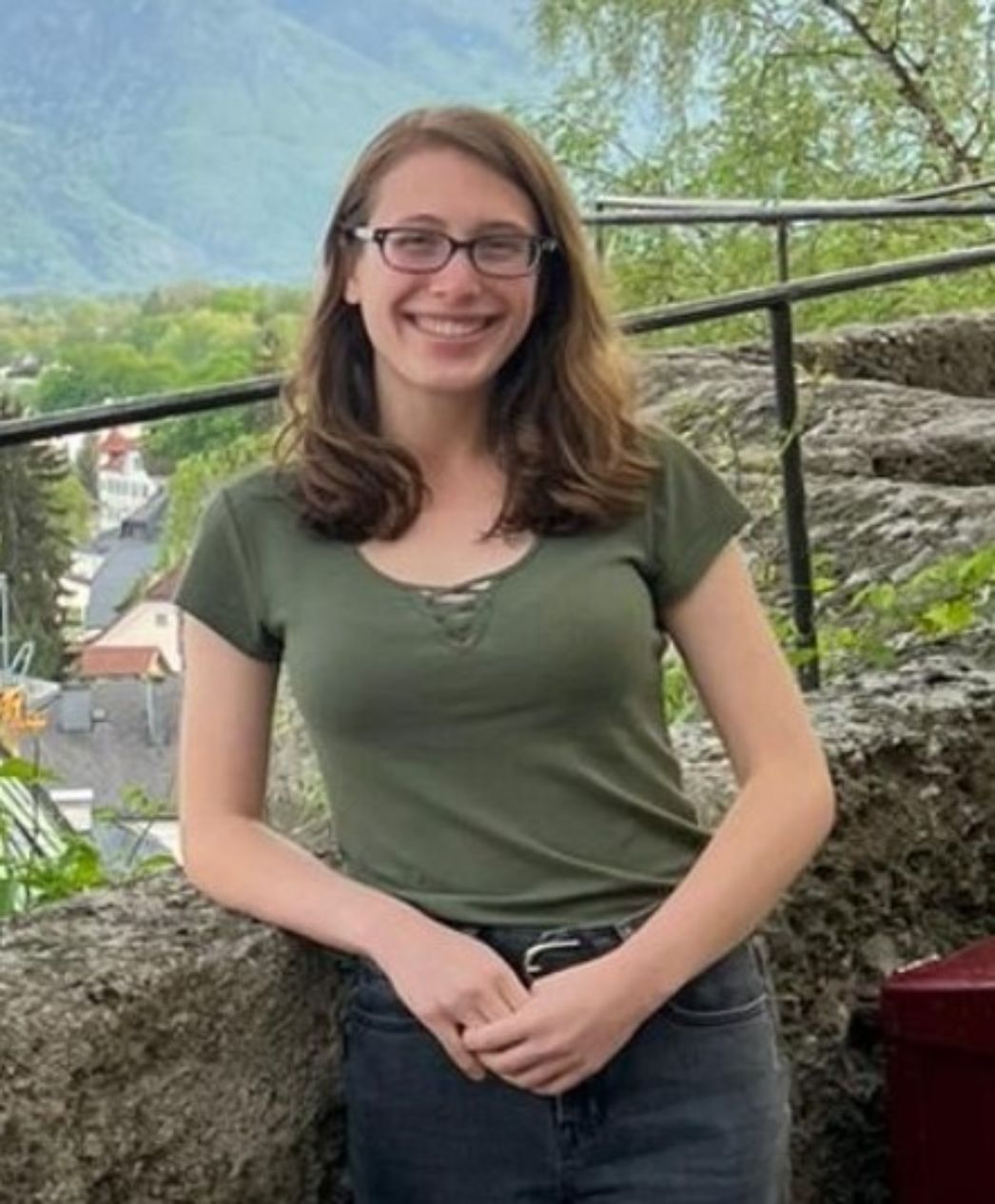 Junge Frau mit schulterlangem braunem Haar und Brille steht draußen vor einer Steinmauer. Sie trägt ein olivgrünes T-Shirt und dunkle Jeans, lächelt freundlich in die Kamera. Im Hintergrund sind grüne Bäume, Häuser und ein bewaldeter Berghang zu sehen.