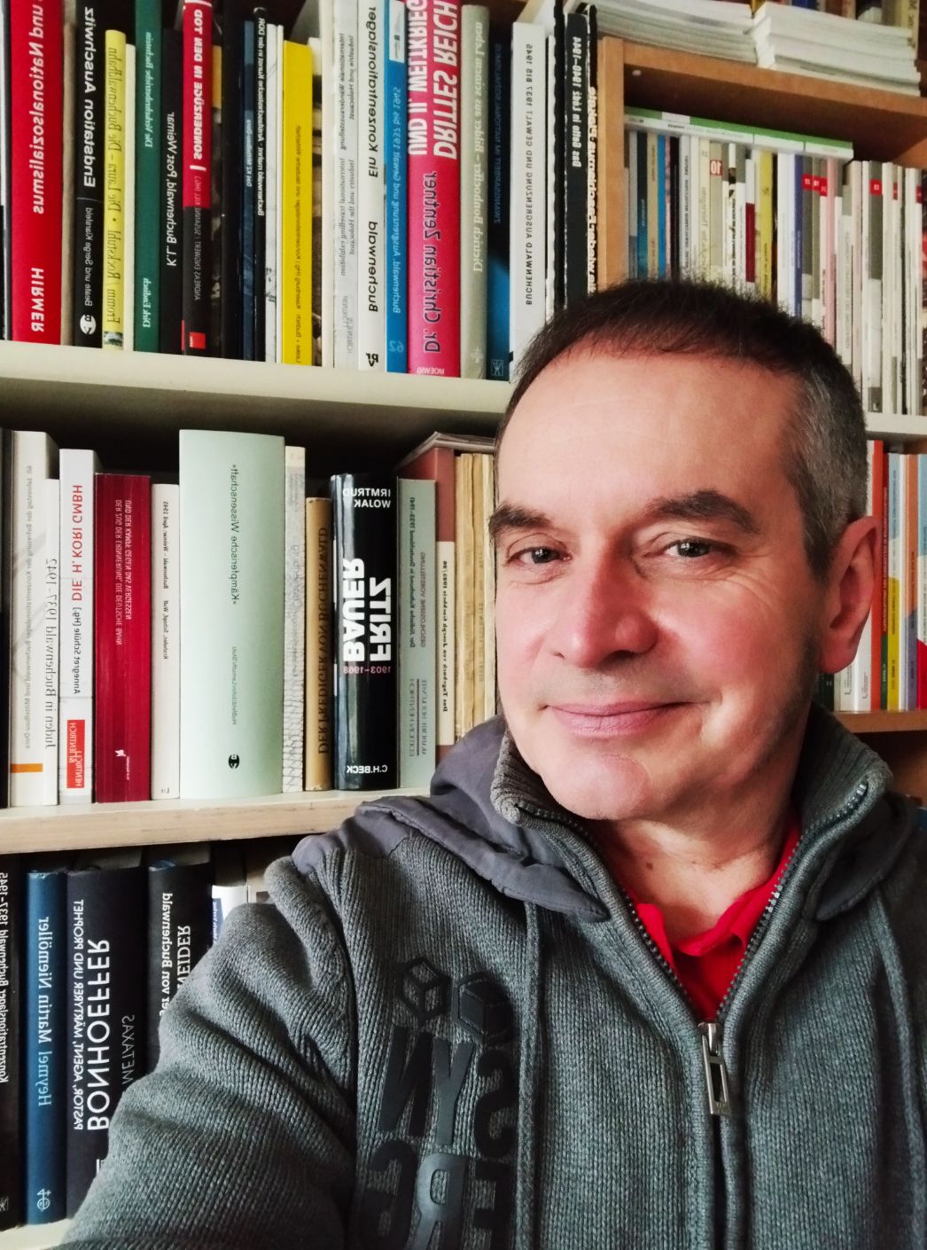Ein Mann mittleren Alters mit kurzen dunklen Haaren lächelt in die Kamera. Er trägt eine graue Kapuzenjacke über einem roten Shirt. Im Hintergrund steht ein gut gefülltes Bücherregal mit Büchern in verschiedenen Sprachen.