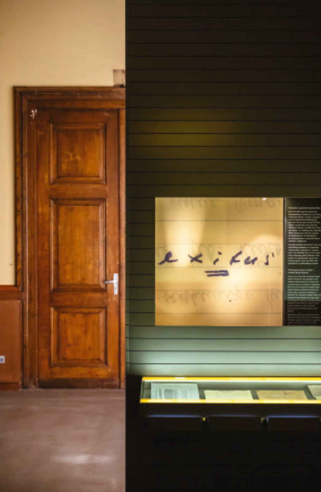 Innenaufnahme eines Ausstellungsraums mit historischer Holzverkleidung und moderner Ausstellungsvitrine. Links ist eine große, braune Holztür zu sehen. Rechts daneben befindet sich ein dunkelgrünes Wandmodul mit einer beleuchteten Vitrine, in der historische Dokumente ausgestellt sind. Darüber ist ein Schriftzug auf hellem Hintergrund zu erkennen, der das Wort ‚exitus‘ zeigt.