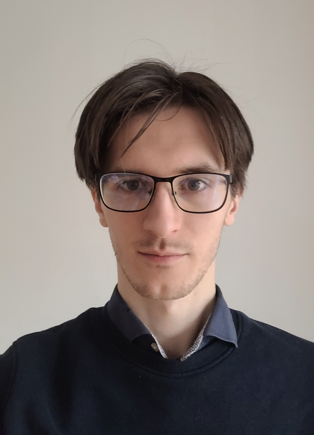 Junger Mann mit kurzen, braunen Haaren und Brille blickt direkt in die Kamera. Er trägt ein dunkelblaues Sweatshirt über einem Hemd mit leicht geöffnetem Kragen. Der Hintergrund ist eine schlichte, helle Wand.