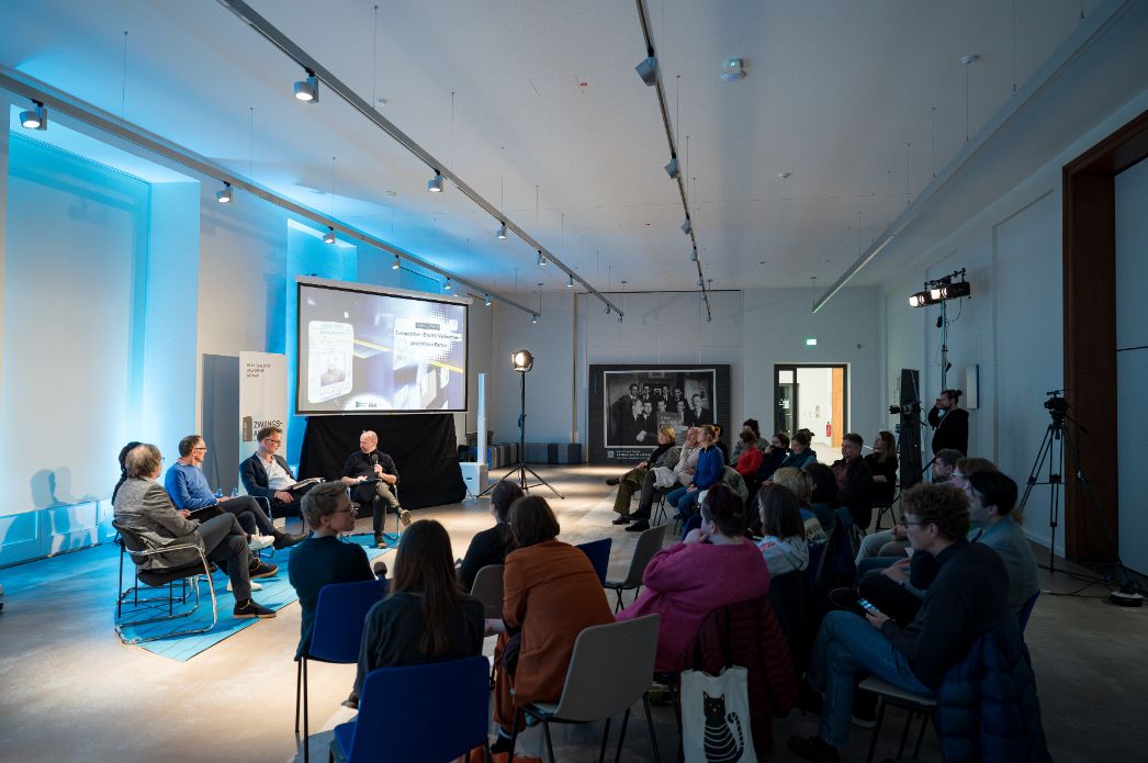 Diskussionsveranstaltung in einem modernen Ausstellungsraum. Vier Personen sitzen auf einem Podium vor einem großen Bildschirm, auf dem ‚Zwangsarbeit. Eine europäische Geschichte‘ zu lesen ist. Im Publikum sitzen zahlreiche Zuhörer:innen. Die Veranstaltung wird professionell gefilmt und beleuchtet.