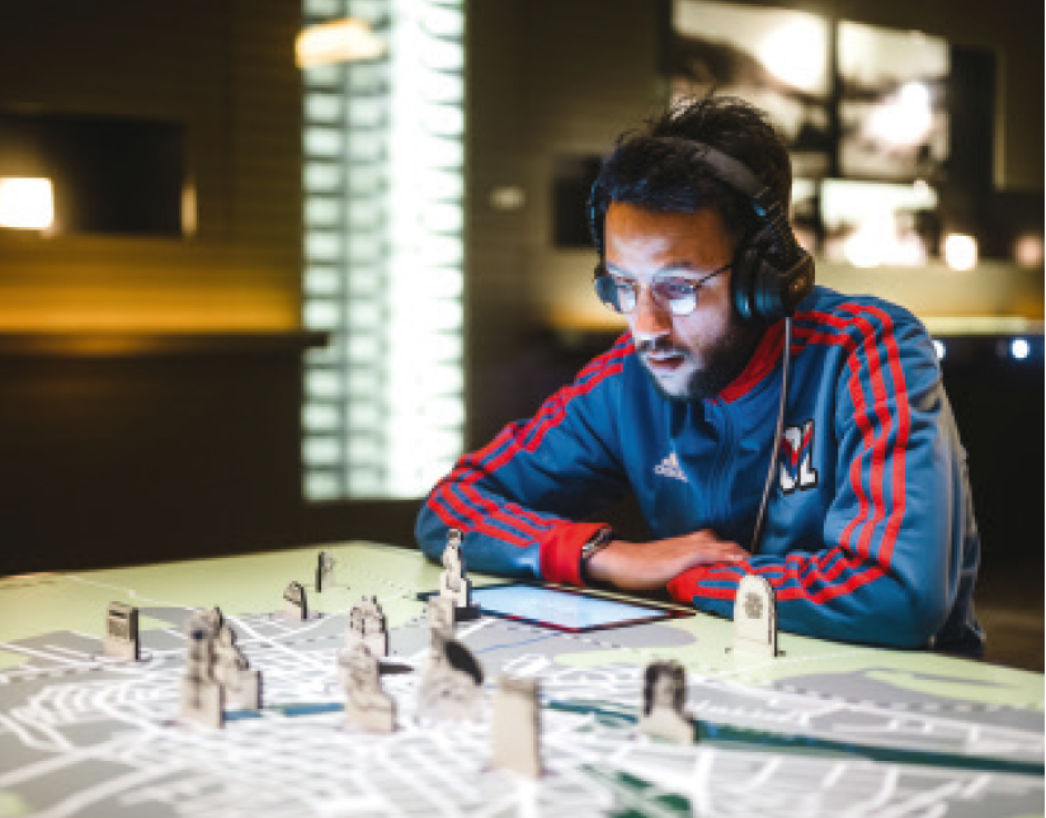 Ein junger Mann mit Kopfhörern sitzt an einem interaktiven Medientisch in einer Ausstellung. Vor ihm befindet sich ein beleuchteter Stadtplan mit aufgestellten Figuren und Gebäuden. Er schaut konzentriert auf ein Tablet oder Display und hört aufmerksam zu.