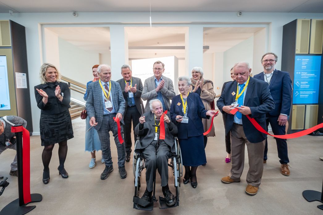 Gruppenfoto einer feierlichen Eröffnung: Im Mittelpunkt sitzen zwei ältere Personen im Rollstuhl, beide mit einer Schere in der Hand, während sie ein rotes Band durchschneiden. Sie werden umringt von weiteren älteren und jüngeren Menschen, die applaudieren und lächeln. Einige tragen Namensschilder und gelbe Bänder, der Ort wirkt modern und hell, vermutlich eine Ausstellung oder ein Museum.