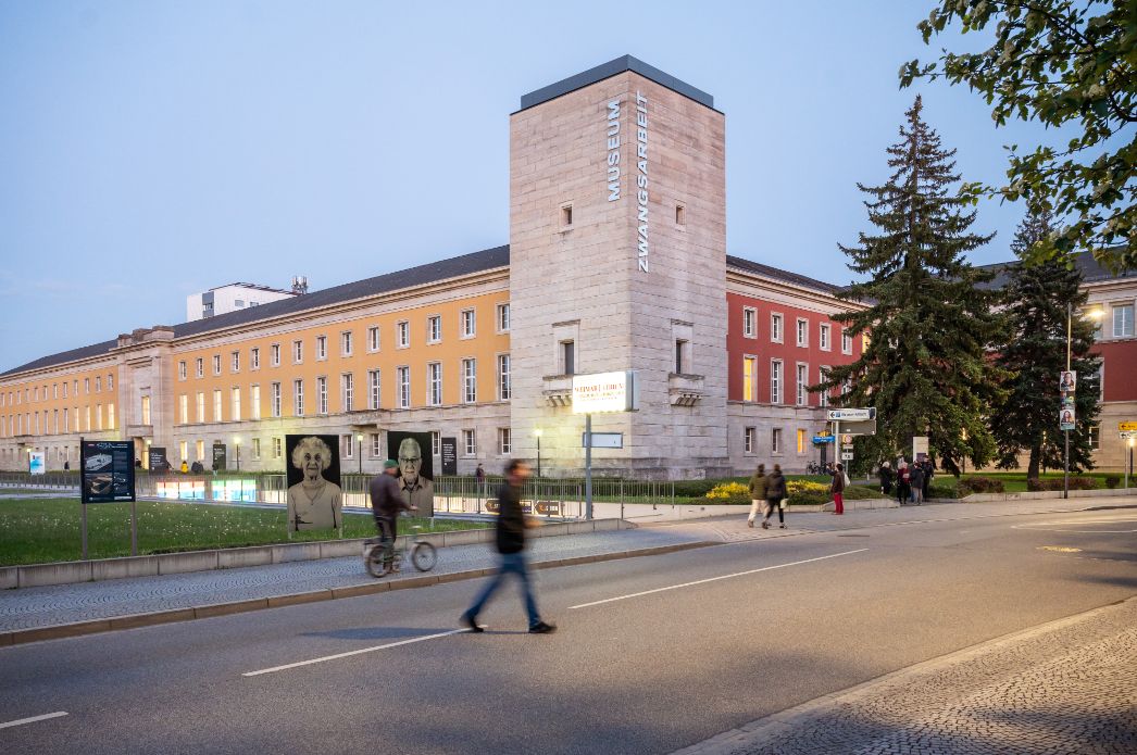 Abendaufnahme des Museums Zwangsarbeit im ehemaligen Haus der Wehrmacht in Berlin. Das historische Gebäude ist in warmen Farbtönen gestrichen, ein markanter Turm trägt den Schriftzug ‚MUSEUM ZWANGSARBEIT‘. Im Vordergrund passieren Fußgänger:innen und ein Radfahrer die Straße. Zwei große Porträtfotos älterer Menschen auf dem Rasen thematisieren Erinnerung und Zeitzeugenschaft.
