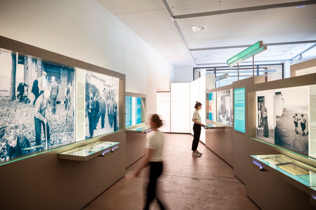 Innenaufnahme einer modernen Ausstellung im Museum zur Zwangsarbeit. Großformatige historische Fotografien und Texte sind hinterleuchtet in Wandvitrinen präsentiert. Zwei junge Frauen betrachten die Exponate; eine bewegt sich unscharf durch den Raum, die andere steht ruhig vor einer Vitrine. Die Atmosphäre ist hell, sachlich und informativ.
