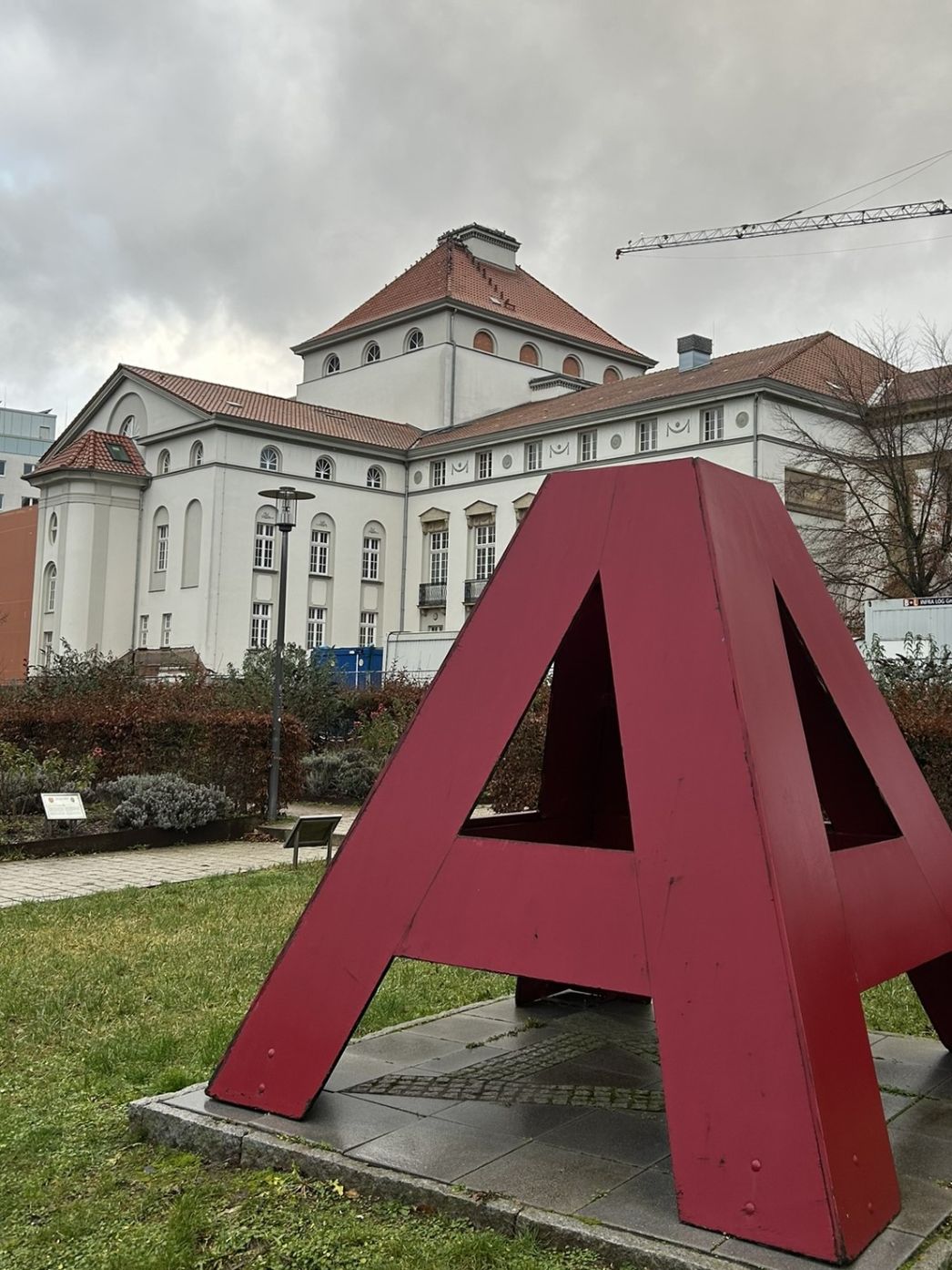 Moderne, rote Stahlskulptur in Form eines überdimensionalen Buchstabens 'A' auf einer kleinen gepflasterten Fläche in einem gepflegten Grünbereich vor einem historischen Gebäude mit hellem Putz und rotem Ziegeldach; im Hintergrund ist ein Baukran zu sehen.