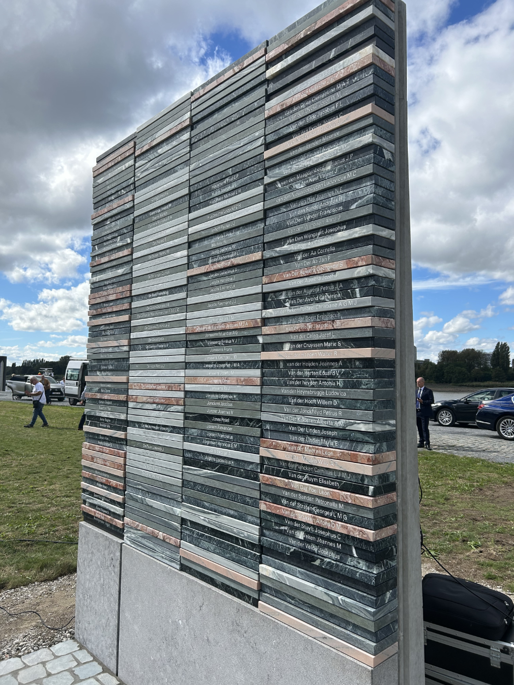Gedenkstele aus gestreiften Steinplatten mit eingravierten Namen, aufgenommen bei Tageslicht unter wolkigem Himmel; im Hintergrund Wiese, Autos und einige stehende Personen.