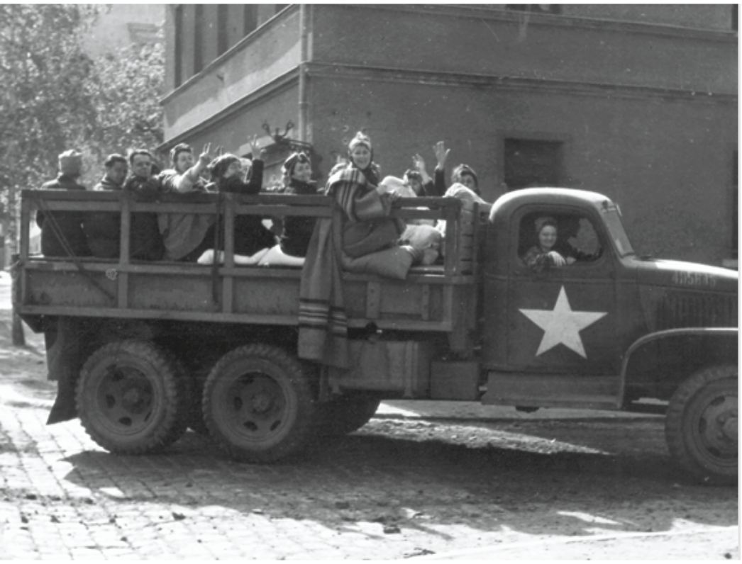 Das Schwarzweißbild zeigt einen offenen Militärlastwagen mit einem großen weißen Stern an der Fahrerkabine – ein Symbol der US-Armee. Auf der Ladefläche sitzen viele Menschen dicht beieinander, in Decken gehüllt, einige winken und lächeln in die Kamera. Es handelt sich vermutlich um befreite KZ-Häftlinge kurz nach der Rettung. Die Stimmung wirkt erleichtert und hoffnungsvoll.