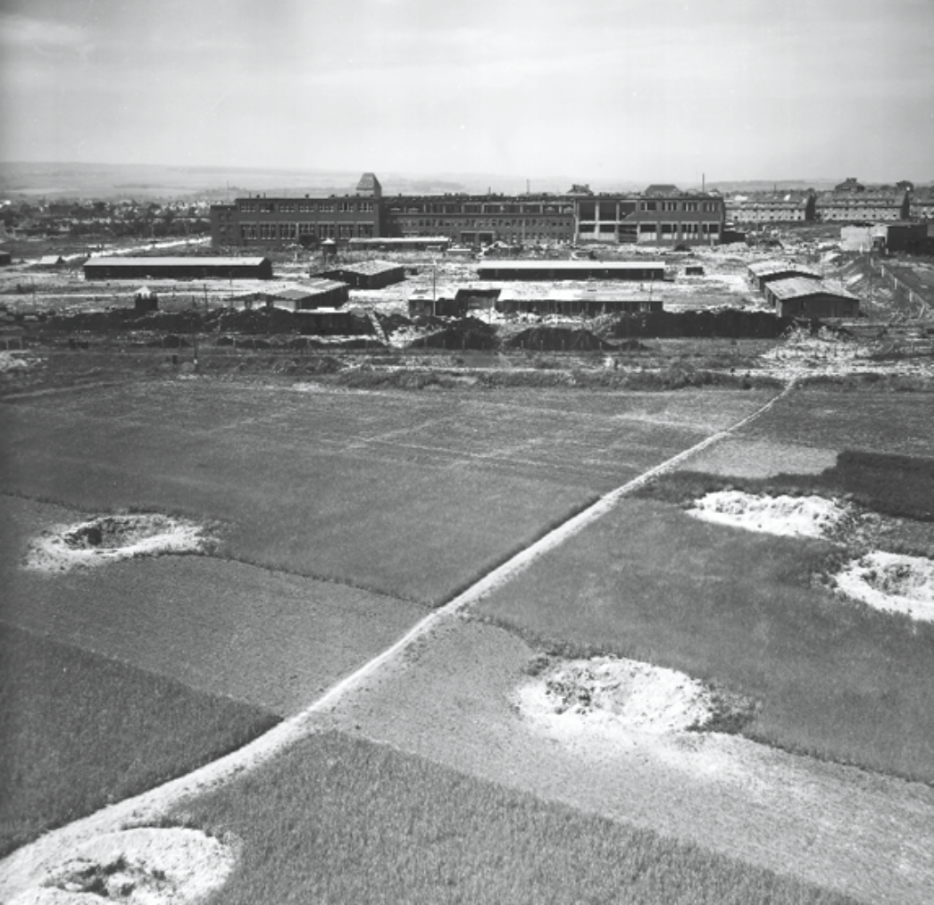 Das Schwarzweißbild zeigt ein weitläufiges Gelände mit Feldern im Vordergrund, durchzogen von Wegen und mehreren großen Bombenkratern. Dahinter liegt ein großes, teilweise zerstörtes Fabrik- oder Lagergebäude mit mehreren Baracken und Umzäunungen. Im Hintergrund erstreckt sich eine Stadtlandschaft. Die Szene wirkt still und verlassen.