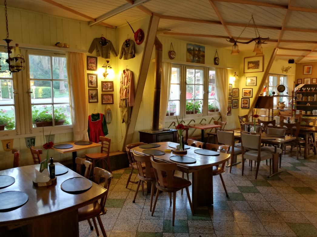 Ein gemütlich eingerichteter Speiseraum mit zahlreichen Holztischen und -stühlen. Die Tische sind mit runden Platzsets gedeckt, auf einigen stehen Blumenvasen mit roten Blüten. An den hellgelben Holzpaneelwänden hängen zahlreiche Bilder, Trophäen und traditionelle deutsche Kleidung, darunter Trachten und Kinderuniformen. Zwei große Fenster mit weißen Vorhängen lassen Tageslicht herein und geben den Blick auf gepflegte Gartenanlagen draußen frei. Die Raumdecke ist mit Holzbalken versehen, von denen ein Geweih mit Lampen hängt. Die Atmosphäre erinnert an ein ländlich-deutsches Gasthaus.