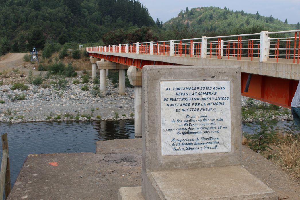 Eine Gedenkplatte aus Stein steht am Ufer eines Flusses in einer bergigen Landschaft mit dichtem Wald. Daneben führt eine moderne Brücke mit weißen und orangen Geländern über den Fluss. Auf der Gedenkplatte ist auf Spanisch zu lesen: „Beim Betrachten dieses Wassers wirst du die Schatten unserer Familien und Freunde sehen, die für das Gedenken an unser Volk segeln.“ Es ist eine Widmung an die Opfer der Diktatur in Colonia Dignidad, deren Leichen in den Fluss Perquilauquén geworfen wurden. Die Gedenktafel stammt von Angehörigenversammlungen der Verschwundenen aus Talca, Linares und Parral.