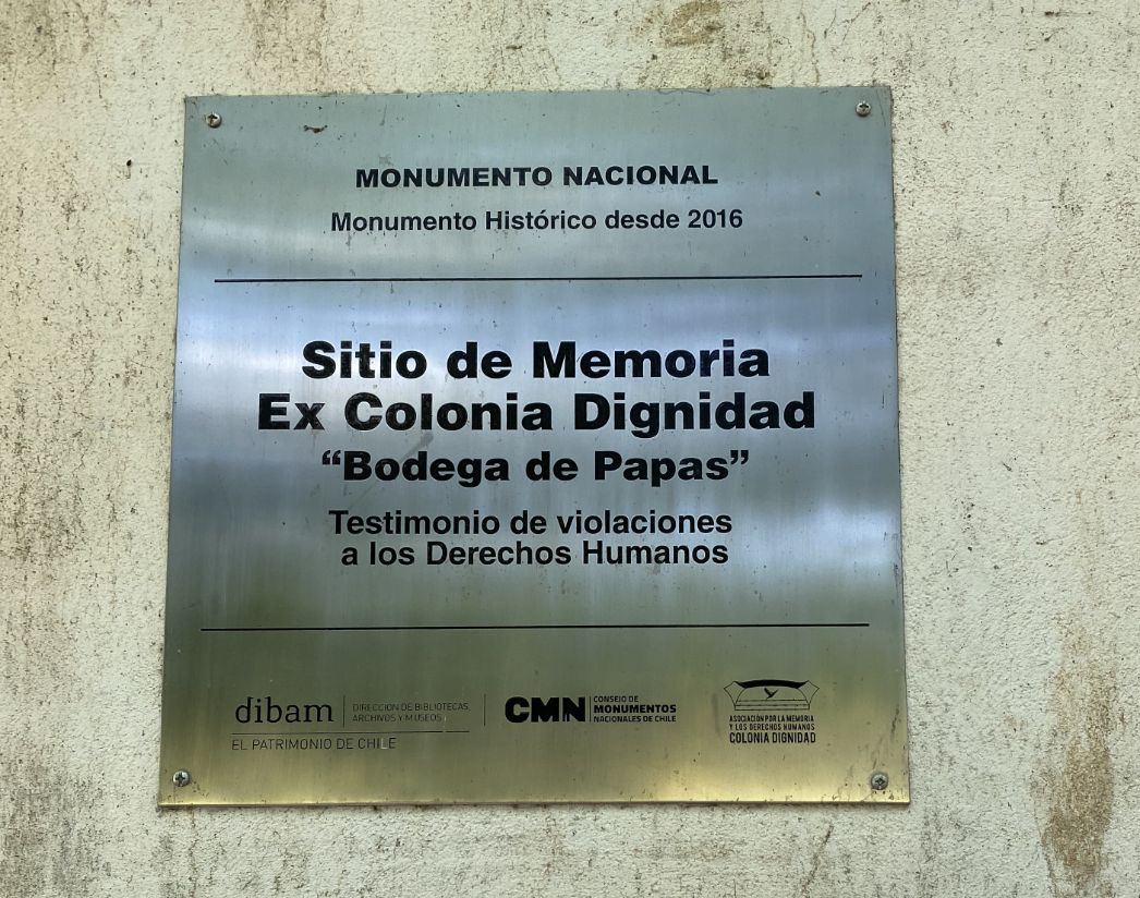 Gedenktafel an einer Wand mit der Aufschrift: „Sitio de Memoria Ex Colonia Dignidad ‘Bodega de Papas’ – Testimonio de violaciones a los Derechos Humanos“. Oben steht: „Monumento Nacional – Monumento Histórico desde 2016“. Unten Logos und Namen staatlicher und zivilgesellschaftlicher Institutionen Chiles, darunter „dibam“, „CMN“ und „Asociación por la Memoria y los Derechos Humanos Colonia Dignidad“.