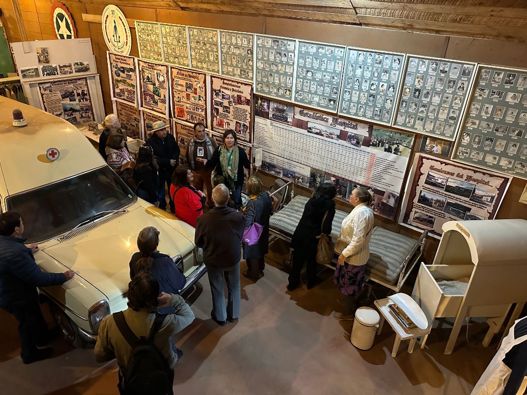 Eine Gruppe von Menschen steht in einem Ausstellungsraum, der an ein kleines Museum erinnert. Links im Bild steht ein altes, cremefarbenes Rettungsfahrzeug mit rotem Kreuz auf dem Dach. An den Wänden hängen zahlreiche Fotos, Plakate und Informationstafeln, die sich offenbar mit der Geschichte eines Krankenhauses oder einer medizinischen Einrichtung befassen. In der Mitte und rechts sind alte Krankenhausbetten, medizinische Geräte und eine Waage ausgestellt. Die Besucher:innen hören einer Person zu, die offenbar eine Führung leitet.