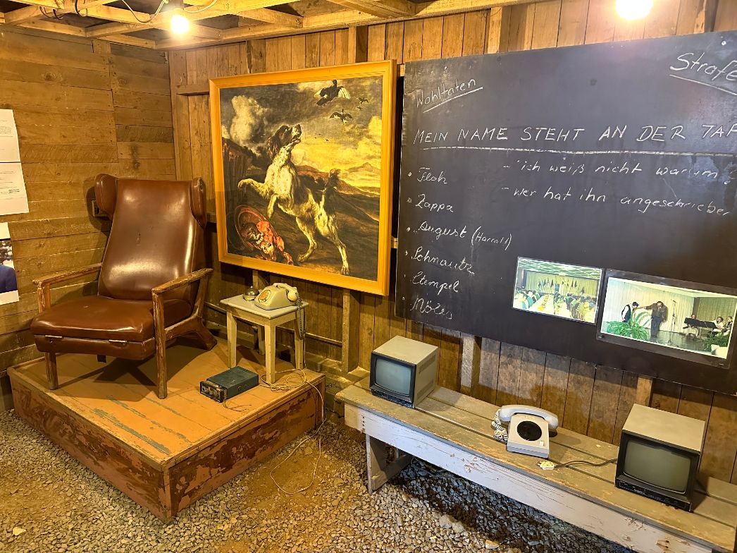 Ein Ausstellungsraum mit Holzverkleidung zeigt eine rekonstruierte Szene aus der Vergangenheit. Links steht ein abgenutzter brauner Ledersessel auf einem Podest, daneben ein kleiner Tisch mit einem alten Telefonapparat und einem Funkgerät. An der Wand hängt ein großes Gemälde mit einem kämpfenden Hund. Rechts davon ist eine Tafel mit Kreidebeschriftung zu sehen, auf der unter den Überschriften „Wohltaten“ und „Strafen“ handschriftlich Namen und Aussagen wie „Mein Name steht an der Tafel“ oder „ich weiß nicht warum“ notiert sind. Darunter stehen zwei kleine Röhrenbildschirme und ein weiteres Telefon. Auf der Tafel sind zwei kleine Farbfotos von einer Versammlung befestigt. Der Boden besteht aus grobem Kies.