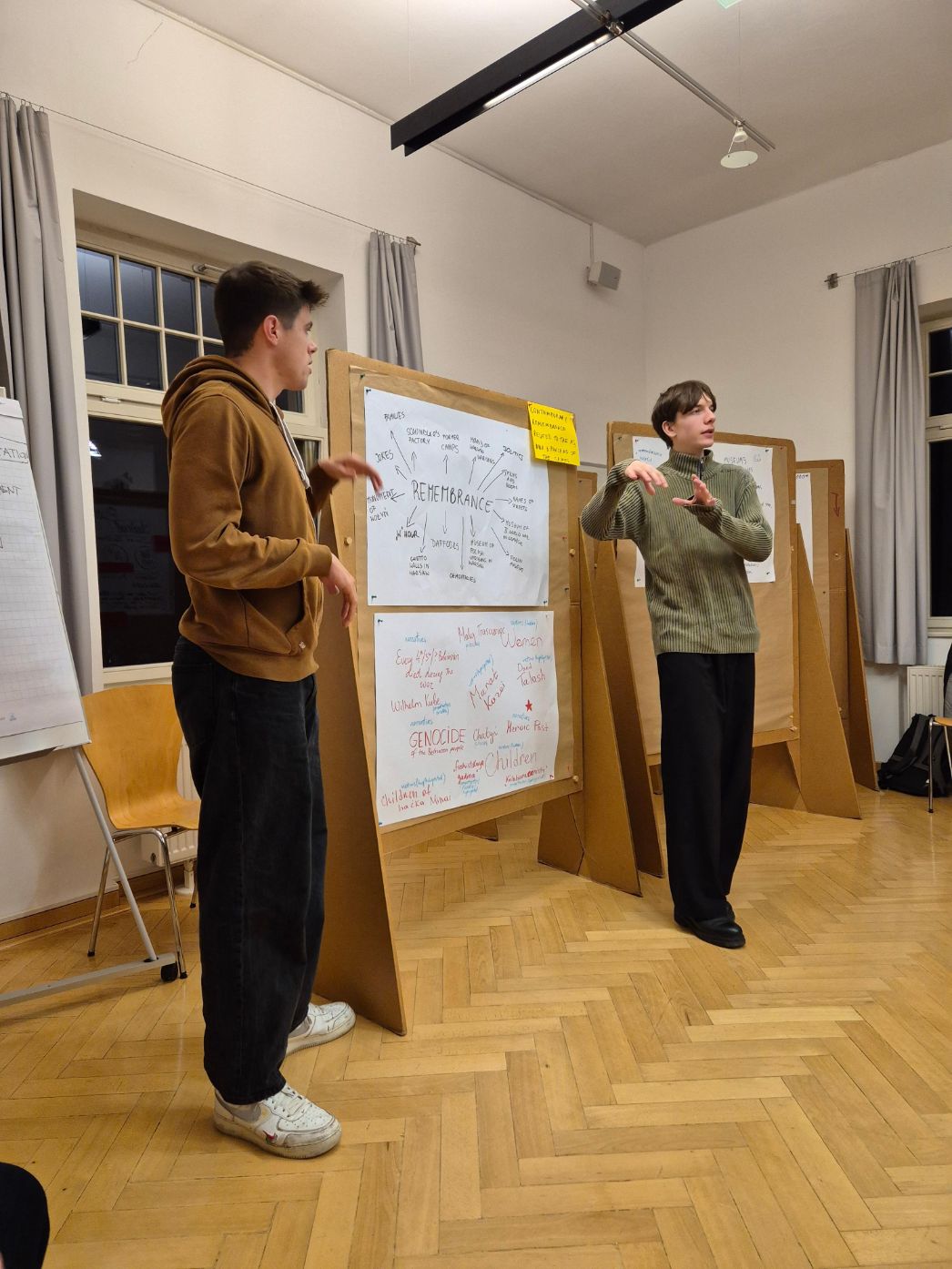 Zwei junge Menschen stehen in einem Seminarraum vor Pinnwänden mit Mindmaps und Stichwörtern zu Erinnerungskultur und Genozid. Sie präsentieren und gestikulieren engagiert. Im Hintergrund sind weitere beschriebene Plakate sichtbar.