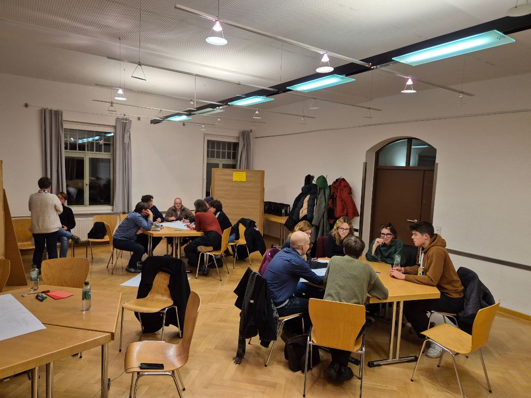 Mehrere Gruppen von Personen sitzen in einem hell erleuchteten Seminarraum an Tischen und diskutieren intensiv. An den Wänden hängen Pinnwände und Jacken. Im Vordergrund steht ein leerer Tisch mit Wasserflaschen und Schreibmaterial.