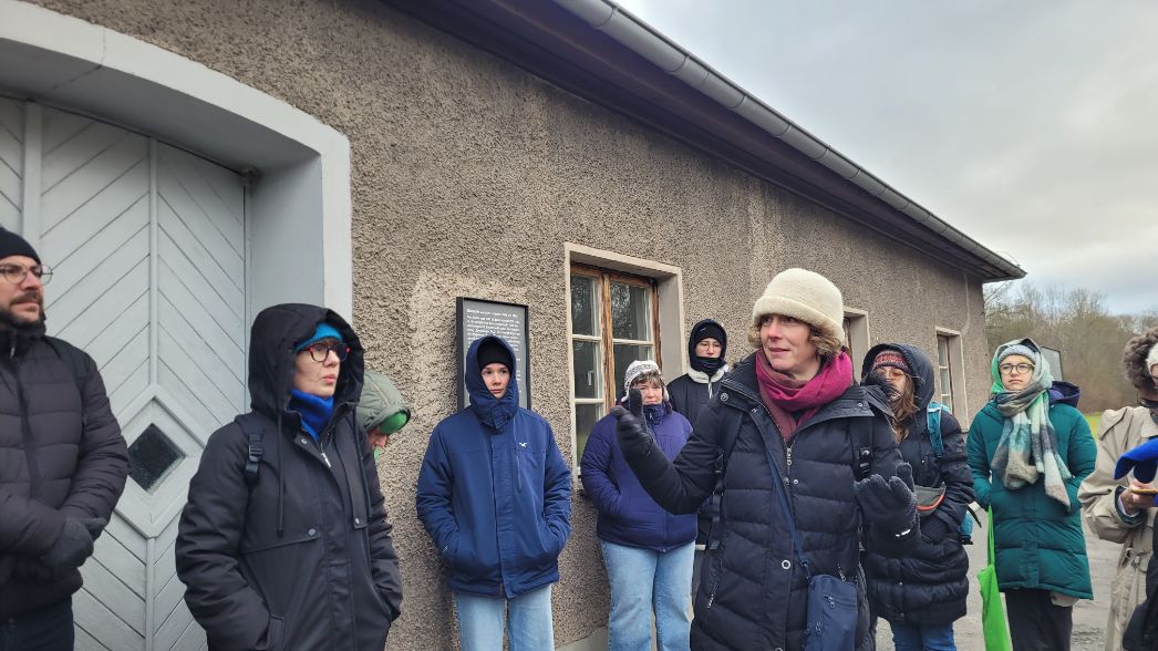 Eine Gruppe von Personen in Winterkleidung steht vor einem historischen Gebäude mit grauer Putzfassade. Eine Frau in der Mitte spricht gestikulierend, während die anderen zuhören. An der Wand hinter ihnen ist ein Informationsschild angebracht.