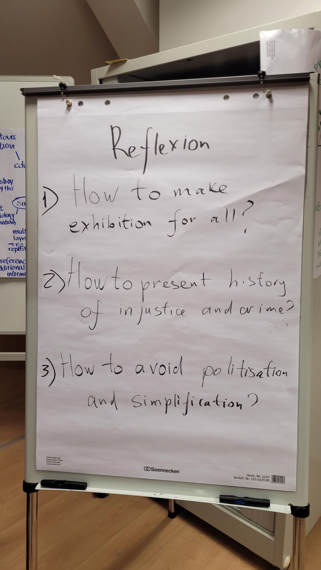 Ein Flipchart mit dem Titel „Reflexion“ zeigt drei handgeschriebene Fragen auf Englisch: How to make exhibition for all? How to present history of injustice and crime? How to avoid politisation and simplification?