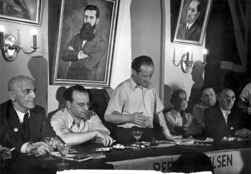 ieses historische Foto zeigt eine bedeutende Szene aus der Nachkriegszeit jüdischer Überlebender, vermutlich aus einer Versammlung der Überlebenden von Bergen-Belsen, einem ehemaligen Konzentrationslager und später Displaced Persons (DP) Camp in der britischen Zone Deutschlands. Im Zentrum des Bildes, stehend und mit Zigarette in der Hand, befindet sich Josef Rosensaft – ein Überlebender des Holocaust und später eine der führenden Stimmen der jüdischen DPs in Deutschland.