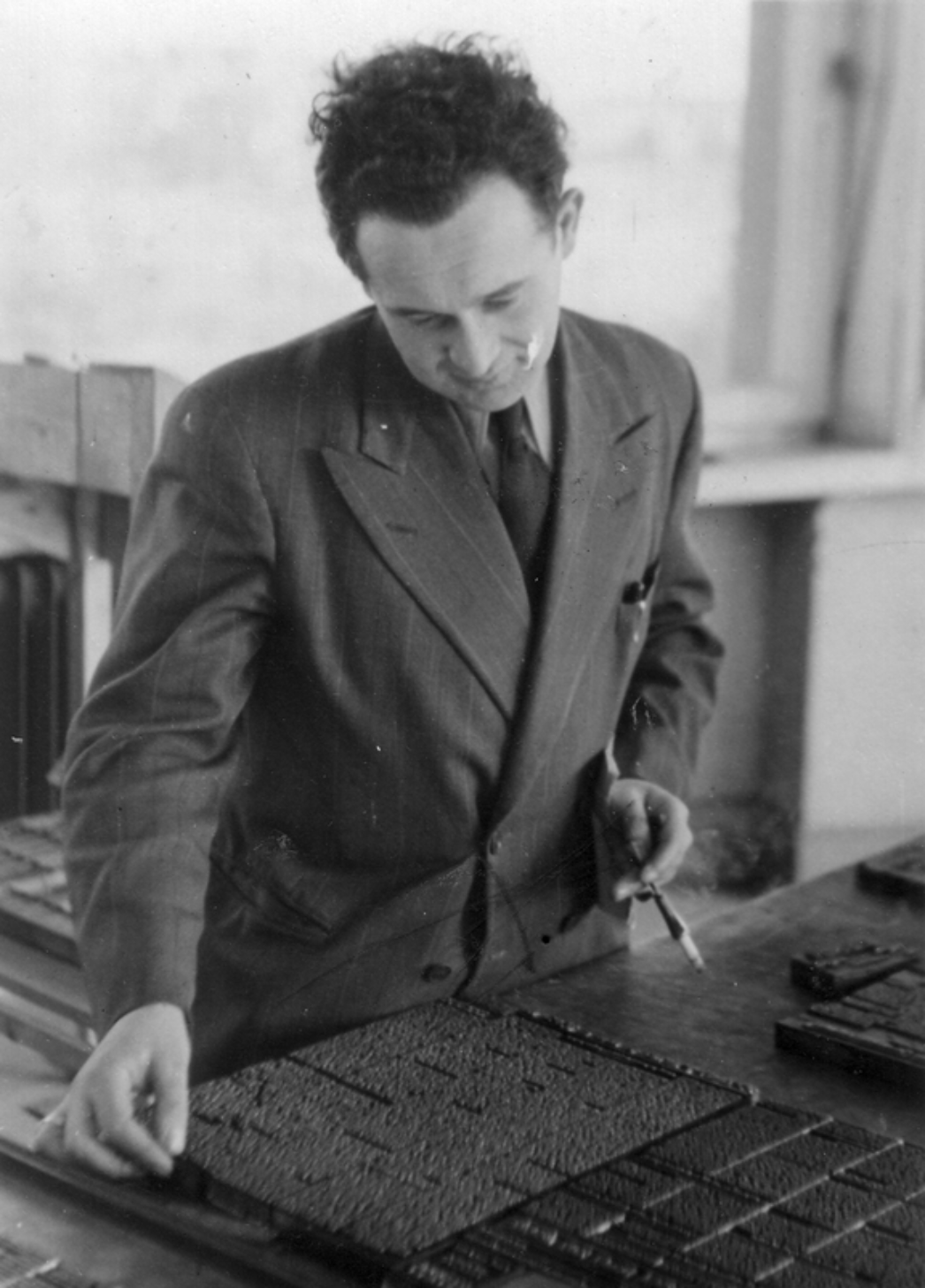 Dieses historische Foto zeigt einen Mann – laut Dateiname handelt es sich vermutlich um Rafael Olewski – bei der Arbeit in einer Druckerei oder Setzerei. Er trägt einen Anzug, hat eine Zigarette im Mundwinkel und hält ein Werkzeug in der Hand (vermutlich eine Pinzette oder ein Setzmesser). Vor ihm auf dem Tisch liegt eine Bleisatzform, also eine Druckform aus einzeln gesetzten Lettern – eine klassische Technik des Buchdrucks, wie sie bis in die Mitte des 20. Jahrhunderts weit verbreitet war.