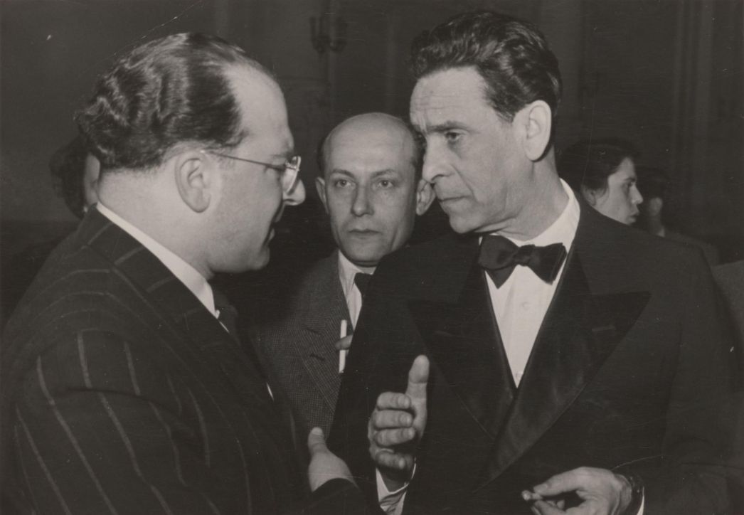 Auf dem Foto sind drei Männer in einem Gespräch zu sehen. Der Mann links trägt einen Nadelstreifenanzug und eine Brille, der Mann rechts einen Smoking mit Fliege und hält eine Zigarette in der Hand. Zwischen den beiden steht ein dritter Mann, der aufmerksam zuhört. Es handelt sich offensichtlich um eine Aufnahme von einem formellen Anlass – möglicherweise eine politische oder gesellschaftliche Veranstaltung, etwa in der Nachkriegszeit.