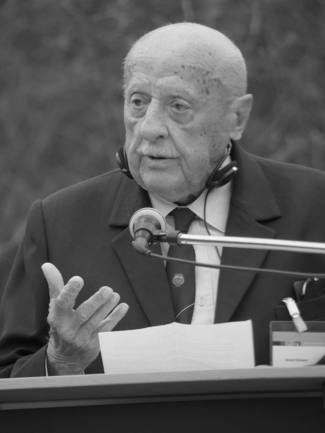 Dieses Foto zeigt den französischen Holocaust-Überlebenden Roland Boisson bei einer Gedenkveranstaltung – vermutlich in einer Gedenkstätte wie Buchenwald oder Mittelbau-Dora. Er steht an einem Rednerpult, spricht in ein Mikrofon und hält eine Rede – möglicherweise auf Französisch, begleitet von Simultanübersetzung (Hinweis: Kopfhörer um den Hals).