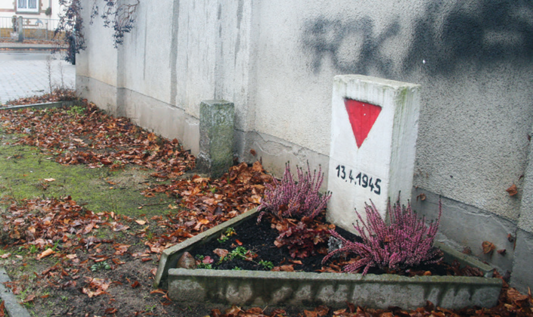 Auf dem Bild ist ein Gedenkstein mit einem roten Dreieck zu sehen. Dieser steht an einer grauen Betonwand mit einem Graffiti "Fuck Nazis".