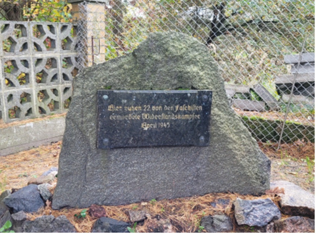 Das Foto zeigt einen großen, dunklen Gedenkstein aus grobem Naturstein. In der Mitte ist eine schwarze Gedenkplatte aus Metall angebracht. Auf dieser steht in deutscher Sprache: „Hier ruhen 22 von den Faschisten ermordete Widerstandskämpfer April 1945“. Der Stein steht im Freien, umgeben von einem Zaun, herbstlichem Laub und ein paar Sitzbänken. Im Hintergrund ist eine Mauer aus Betonsteinen zu sehen.