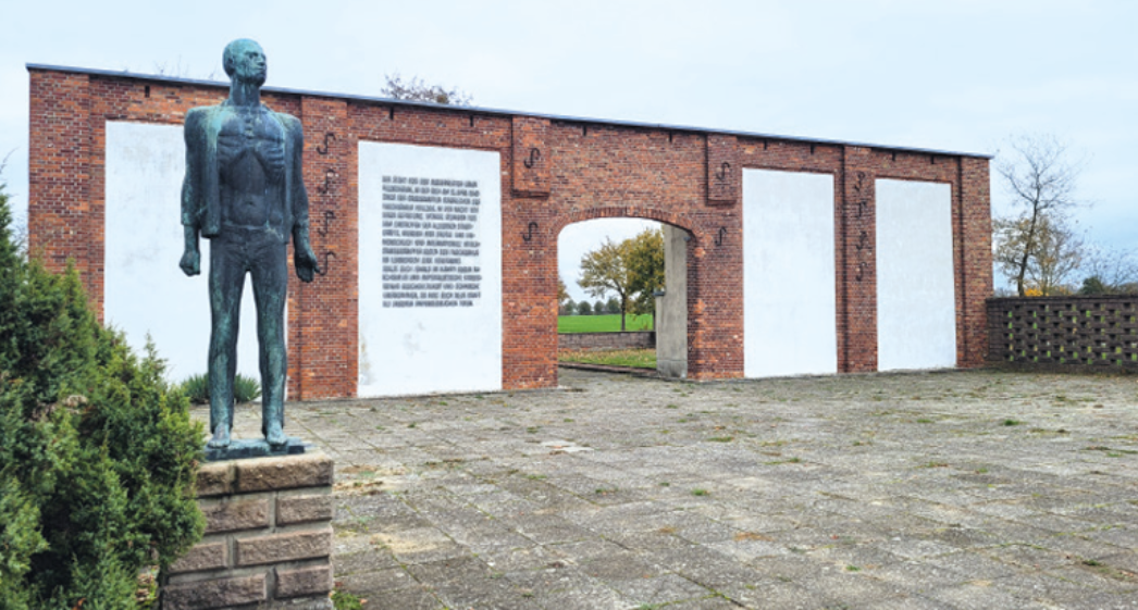 Das Foto zeigt eine Gedenkstätte mit einer roten Backsteinmauer, in die mehrere weiße Gedenktafeln eingelassen sind. In der Mitte befindet sich ein Torbogen, durch den man auf eine Wiese mit Bäumen blickt. Links im Vordergrund steht eine Bronzestatue eines abgemagerten, aufrecht stehenden Mannes mit eingefallenen Gesichtszügen – ein symbolisches Mahnmal für die Leiden der KZ-Häftlinge. Die Fläche vor der Mauer ist gepflastert und weitläufig.