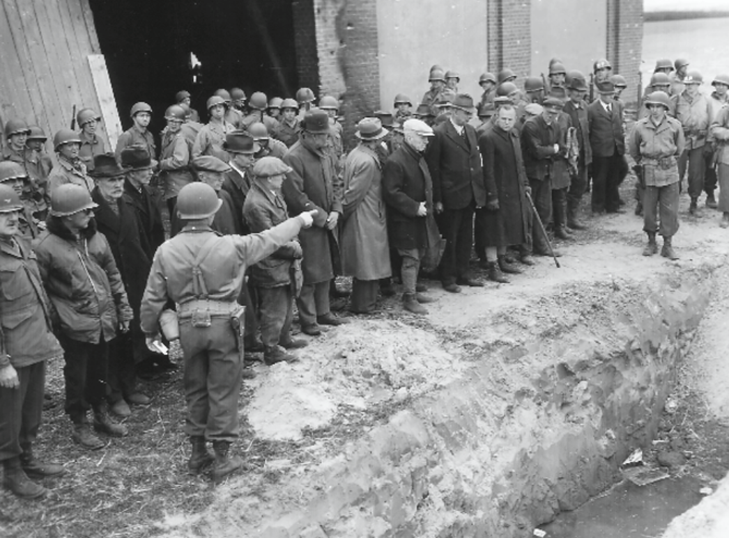 Das Schwarzweißbild zeigt amerikanische Soldaten, die eine Gruppe deutscher Zivilisten an einem offenen Massengrab konfrontieren. Ein Soldat weist in die Grube, während die Zivilisten ernst und betroffen schauen.