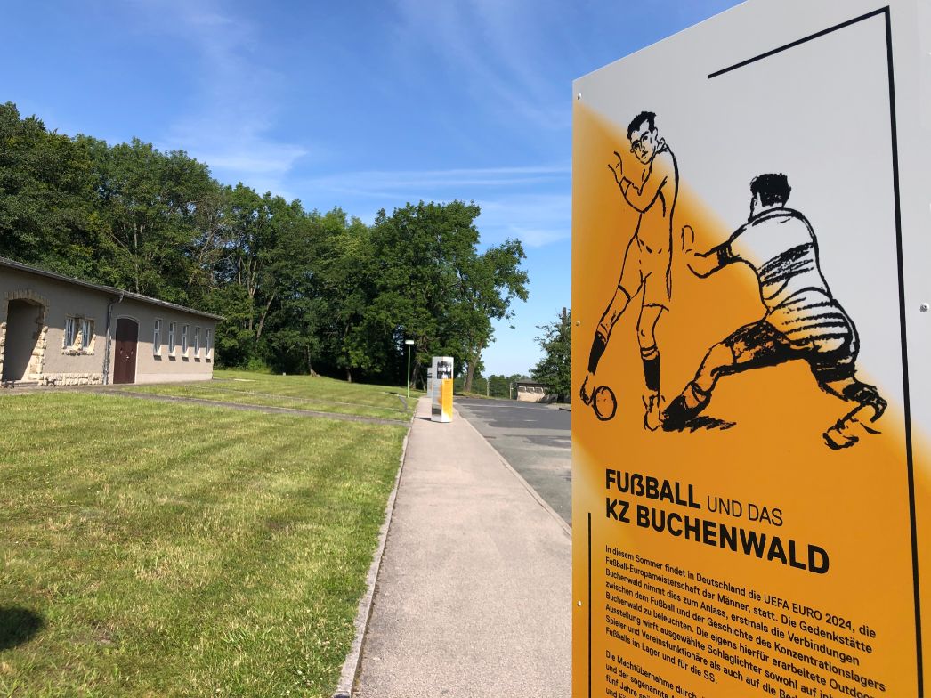 Informationsstele mit Illustration zweier Fußballspieler und der Überschrift „Fußball und das KZ Buchenwald“ auf dem Gelände der Gedenkstätte Buchenwald. Im Hintergrund sind weitere Tafeln sowie ein Gebäude mit hellem Putz und Steineinfassung zu sehen. Der Himmel ist klar und blau, das Gelände gepflegt.