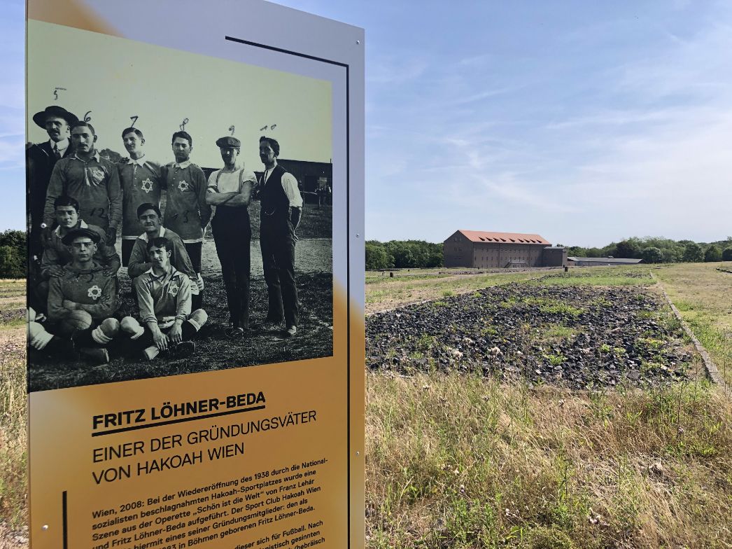 Informationsstele auf dem Gelände der Gedenkstätte Buchenwald mit historischem Foto einer jüdischen Fußballmannschaft, begleitet von einem Text über Fritz Löhner-Beda, einen der Gründungsväter des Vereins Hakoah Wien. Im Hintergrund ist ein langgestrecktes Gebäude mit rotem Satteldach zu sehen, das sich am Rande eines weiten, mit Gras und Schotter bedeckten Geländes befindet. Der Himmel ist blau und klar.