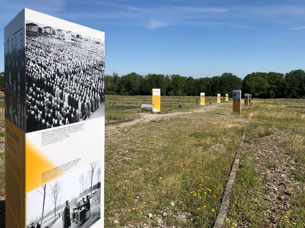 Reihe von Informationsstelen auf dem Gelände des Konzentrationslagers Buchenwald bei blauem Himmel und sonnigem Wetter. Die im Vordergrund stehende Stele zeigt historische Schwarz-Weiß-Fotografien mit einer großen Gruppe von Häftlingen sowie einer Selektion durch SS-Personal. Weitere gelb-weiße Tafeln sind in der Tiefe entlang eines Weges angeordnet. Umgeben ist die Szenerie von Gras, Blumen, Schotterresten und Wald am Horizont.