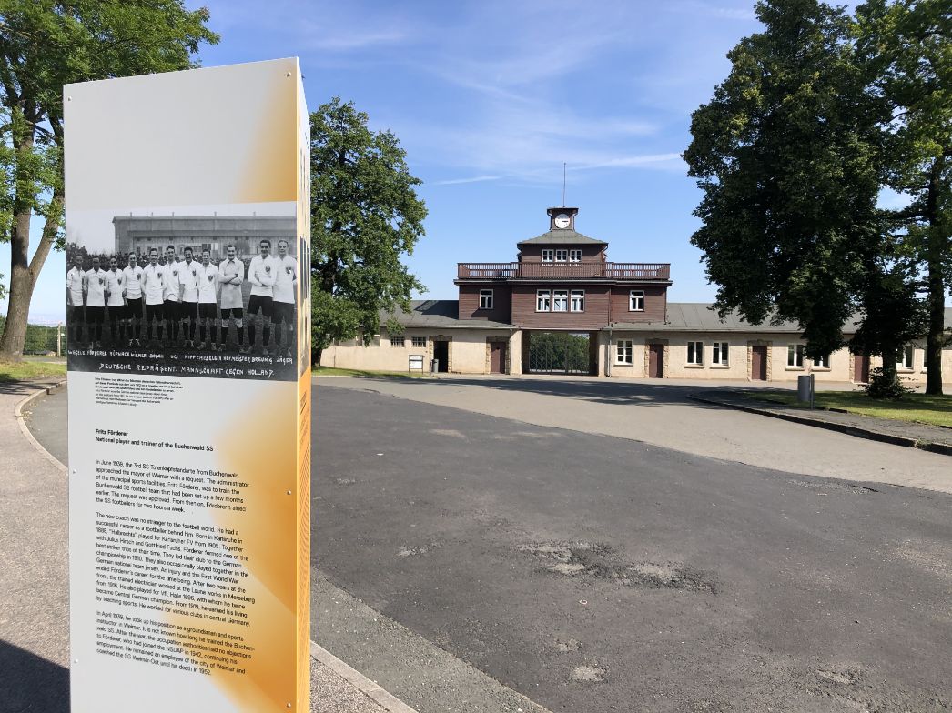 Infotafel über Fritz Förderer, einen ehemaligen Fußballnationalspieler und Trainer der Buchenwald-SS, vor dem historischen Lagertor mit Uhrenturm des Konzentrationslagers Buchenwald. Die Stele zeigt ein historisches Schwarz-Weiß-Foto einer Fußballmannschaft sowie begleitenden Text. Im Hintergrund das ikonische Lagertor mit den Aufschriften „Jedem das Seine“, flankiert von Bäumen und unter blauem Himmel.