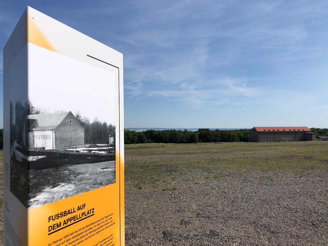 Ein Informationsstele auf einem weiten, offenen Platz unter blauem Himmel. Auf der Stele ist ein historisches Schwarz-Weiß-Foto mit der Überschrift „Fußball auf dem Appellplatz“ zu sehen. Die Stele enthält Textinformationen zur Nutzung des Platzes im Konzentrationslager Buchenwald. Im Hintergrund ist ein großes Gebäude mit rotem Ziegeldach zu erkennen, dahinter ein bewaldeter Horizont. Die Szenerie wirkt ruhig und klar, in starkem Kontrast zur historischen Bedeutung des Ortes.