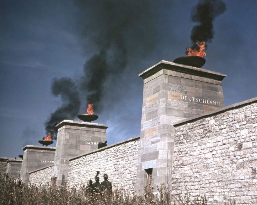 Farbfotografie von Gedenksäulen aus Stein mit brennenden Feuerschalen auf dem Gelände der Gedenkstätte Buchenwald. Die Säulen tragen Länderschriftzüge wie „DEUTSCHLAND“, „DANMARK“ und weitere. Schwarzer Rauch steigt in den blauen Himmel, im Vordergrund sind silhouettenhafte Besucher zu erkennen.
