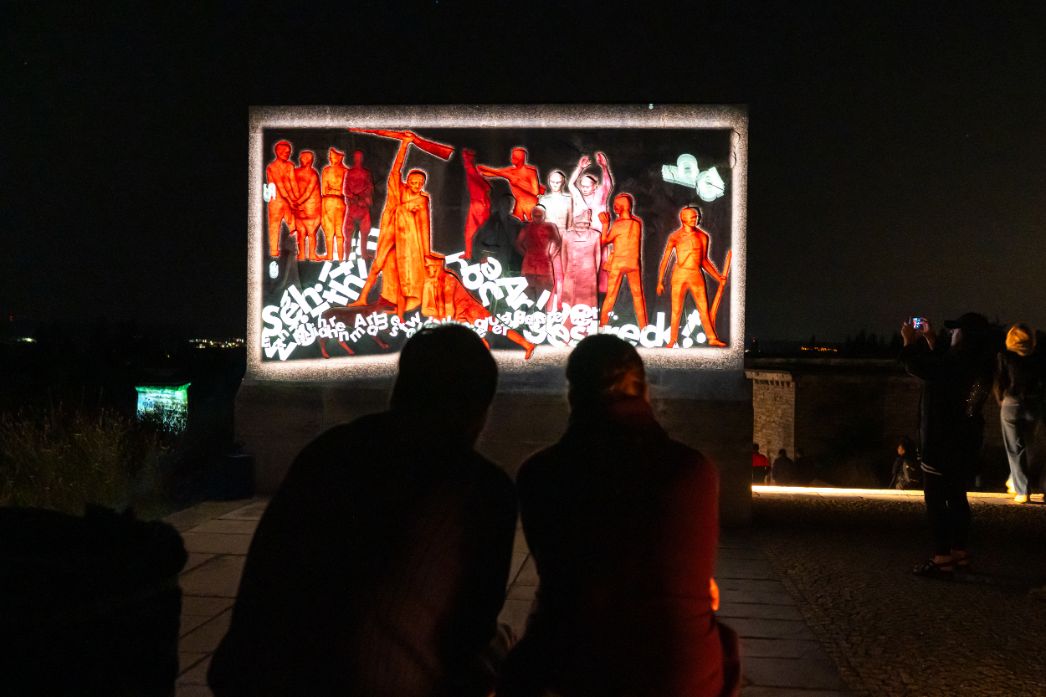 Zwei Personen sitzen nachts vor einem beleuchteten Denkmalrelief mit roten Figuren, auf das Textfragmente projiziert werden. Weitere Menschen stehen im Hintergrund und betrachten oder fotografieren die Projektion.