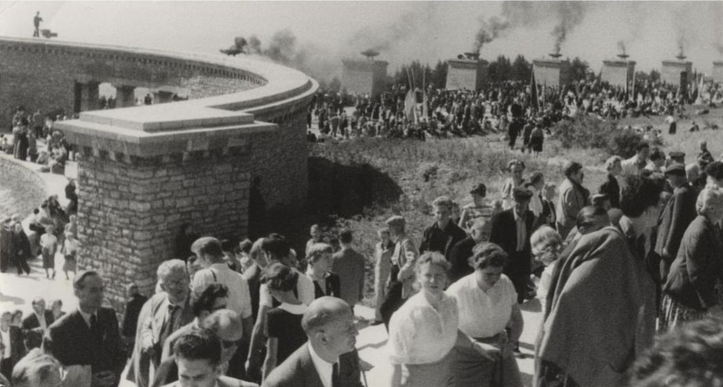 Schwarz-Weiß-Fotografie einer großen Menschenmenge bei einer Gedenkveranstaltung. Im Vordergrund steigen Menschen eine breite Treppe hinauf. Im Hintergrund sind viele Personen auf einem Feld vor monumentalen Steinsäulen mit brennenden Feuerschalen zu sehen.