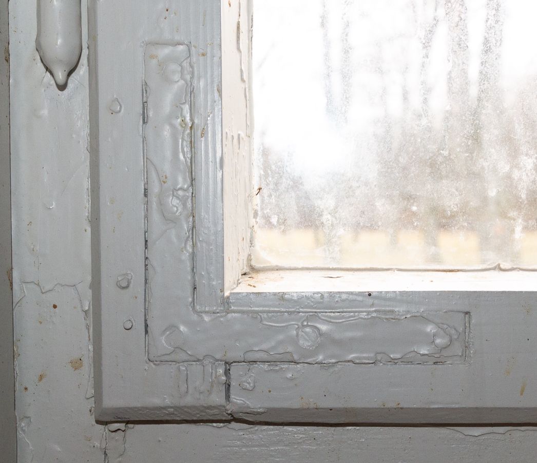 Detailaufnahme einer stark überstrichenen Fensterinnenecke. Sichtbar ist eine L-förmige Metallverstärkung auf dem hölzernen Fensterrahmen, die mehrmals übermalt wurde. Die Schraubenköpfe unter der Farbschicht sind erkennbar. Das Fenster ist einfach verglast, die Scheibe wirkt gealtert und ist beschlagen oder verschmutzt. Im Hintergrund sind unscharf Bäume zu sehen.