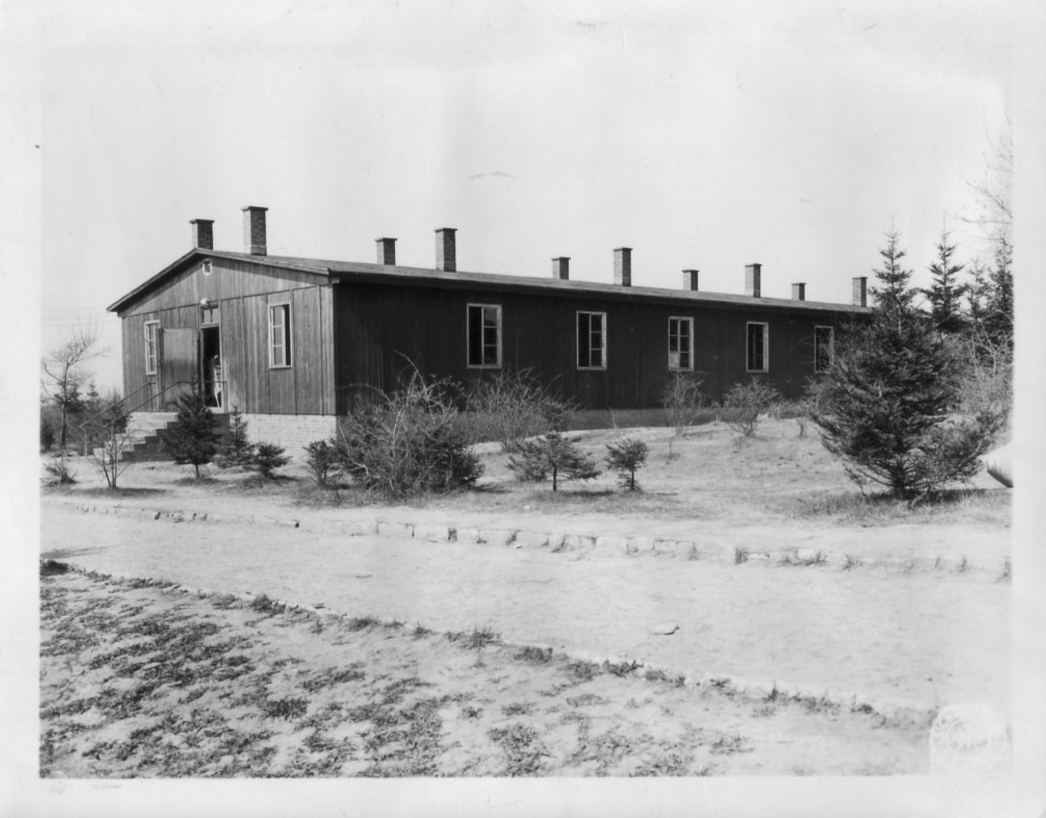 Schwarz-weiß-Fotografie einer langen Holzbaracke mit mehreren Schornsteinen auf dem Satteldach. Die Fassade besteht aus senkrecht verlaufenden Holzbrettern, auf einem gemauerten Sockel stehend. Links ist ein Eingangsbereich mit Treppe und geöffneter Tür zu sehen. Entlang der Längsseite befinden sich regelmäßig angeordnete Fenster. Vor der Baracke wächst spärliche Vegetation mit jungen Nadelbäumen und niedrigen Büschen. Ein unbefestigter Weg führt an der Baracke vorbei, im Hintergrund sind weitere Bäume sichtbar.