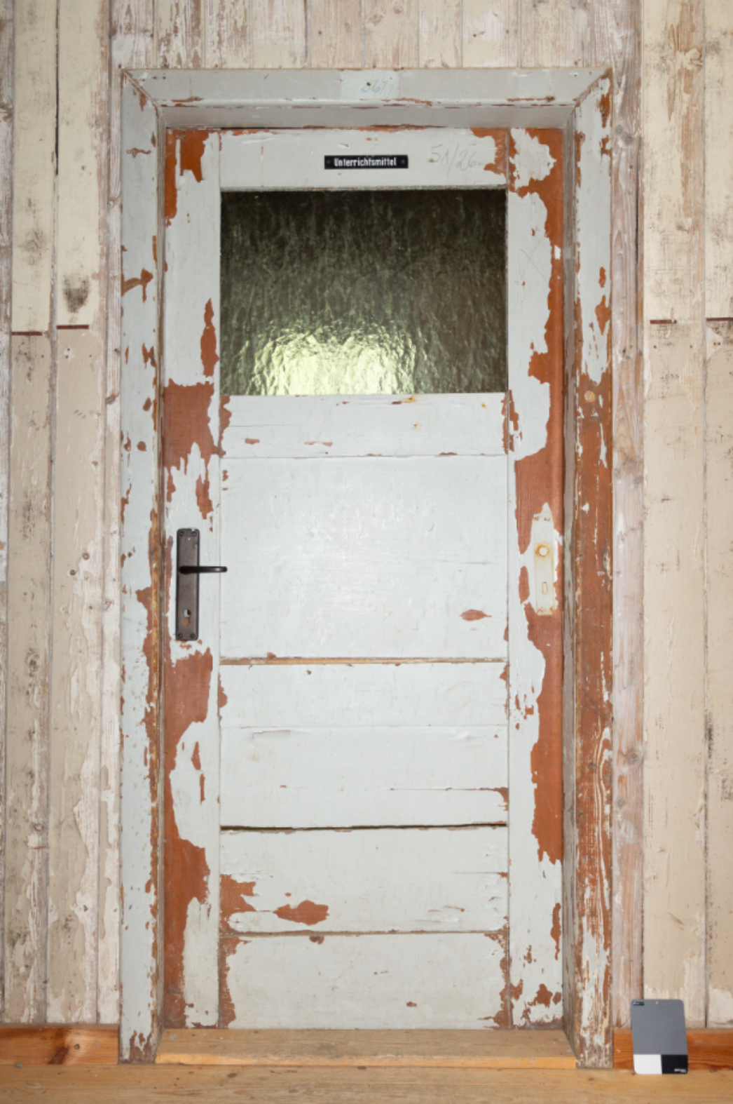 Abgenutzte Holztür mit oberem Fensterbereich aus strukturiertem Glas, eingefasst in einen ebenfalls stark abgeplatzten Türrahmen. Über dem Fenster ist ein schwarzes Schild mit weißer Aufschrift „Unterrichtsmittel“ angebracht. Rechts über dem Türrahmen handschriftlich die Nummer „51126“. Die Tür ist grau gestrichen, mit großflächig sichtbarer rotbrauner Grundierung. Links befindet sich ein schwarzer Türgriff, rechts ein heller Lichtschalter. Unten rechts steht ein Farbkalibrierungstool auf dem Holzboden. Die Wandverkleidung besteht aus verwitterten Holzlatten.