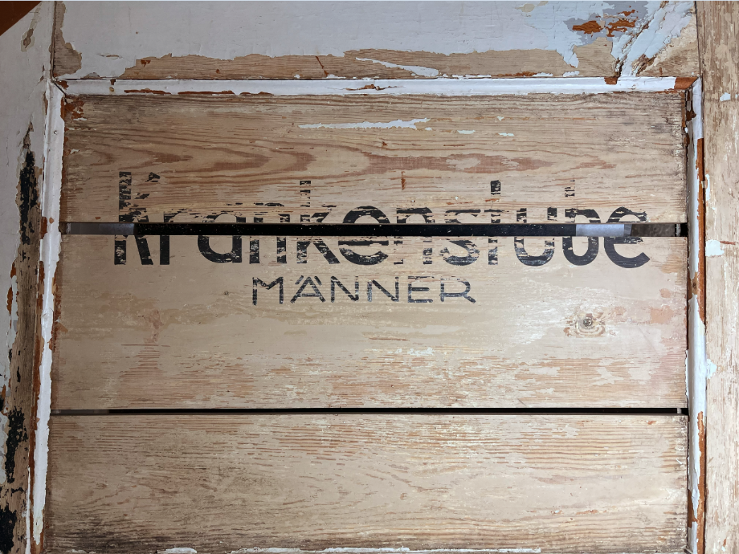 Verwittertes Holzschild mit der schwarzen Aufschrift „Krankenstube Männer“. Die Farbe ist teils abgeblättert, das Schild ist in eine hölzerne Wand eingelassen und weist Spuren von Alterung und Abnutzung auf.