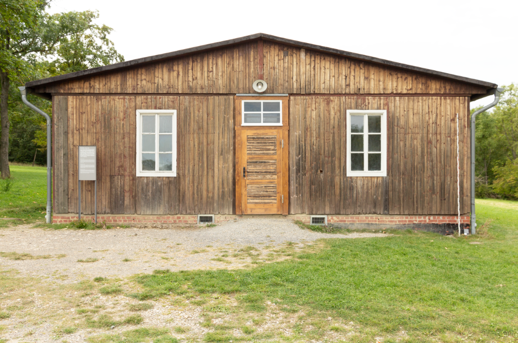 Frontansicht eines schlichten Holzgebäudes mit symmetrischem Aufbau. In der Mitte befindet sich eine Holztür mit Fenster darüber, flankiert von zwei weißen Sprossenfenstern. Das Gebäude steht auf einem Fundament aus Ziegelsteinen, davor ein Schotterweg und Rasen. Links neben dem Fenster ist eine Infotafel aufgestellt.