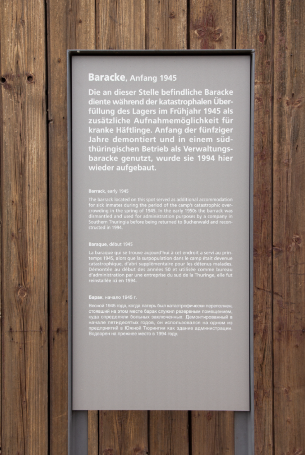 Informationstafel an einer Holzfassade mit viersprachiger Beschriftung (Deutsch, Englisch, Französisch, Russisch). Der deutsche Text beschreibt die Geschichte einer Baracke, die 1945 zur Unterbringung kranker Häftlinge diente, in den 1950er Jahren als Verwaltungsbaracke genutzt und 1994 an diesem Ort wieder aufgebaut wurde.
