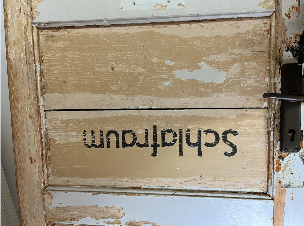 Alte, abgeblätterte Holztür mit der schwarzen Aufschrift „Schlafraum“, die auf dem Bild auf dem Kopf steht. Die Tür zeigt deutliche Gebrauchsspuren, Abnutzung der Farbe und einen Metalltürgriff auf der rechten Seite.