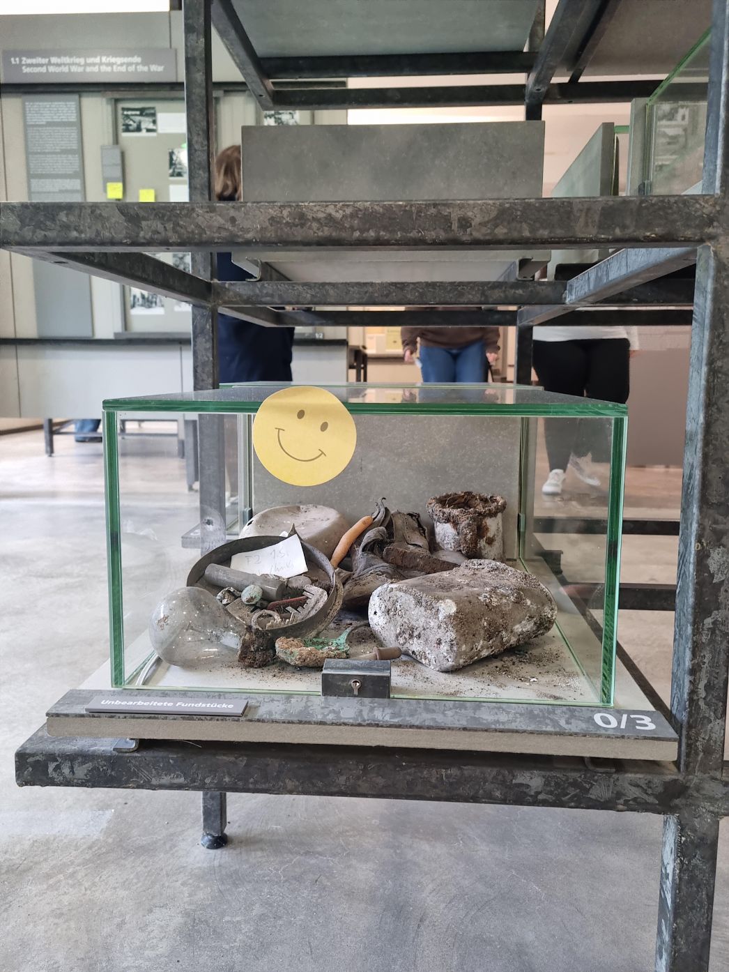 Eine Glasvitrine mit der Beschriftung „Unbearbeitete Fundstücke“ zeigt eine Sammlung von verschmutzten Objekten, darunter ein Schuh, Metallteile, eine Glühbirne und Ziegelreste. Auf der Glasvitrine ist ein gelber Aufkleber mit einem gezeichneten Smiley angebracht. Im Hintergrund sind Ausstellungswände mit Texten und Fotos sowie Besucher:innen der Ausstellung zu sehen.